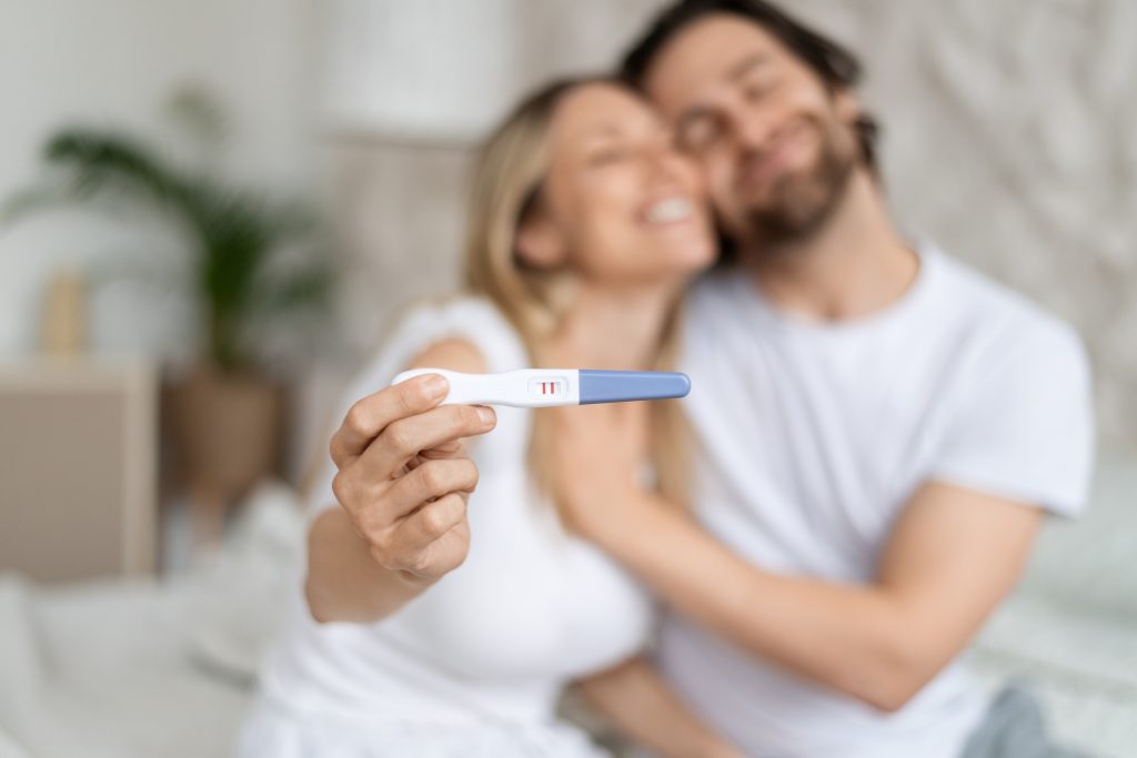 Happy caucasian spouses showing positive pregnancy test and embracing at home, selective focus